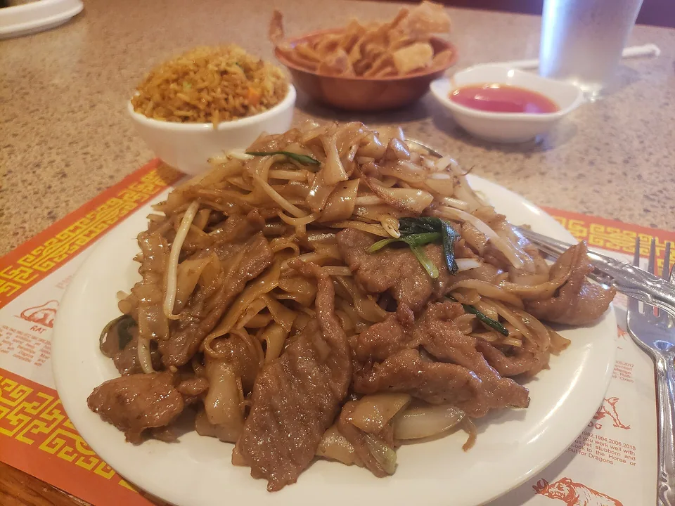 Real finished-dish brief: a dark oval plate of glossy, intact wide noodles with browned beef ribbons, crisp pale bean sprouts, ginger slivers, and scallion sections visibly distributed; no gravy pooling underneath.