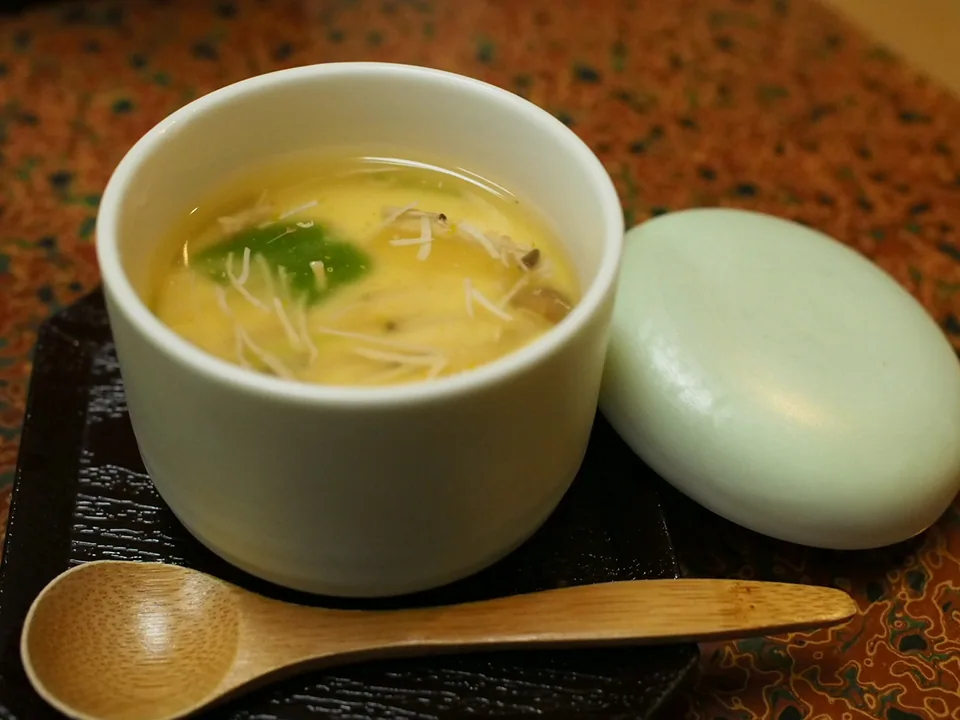 Real photograph of a lidded chawanmushi cup with smooth pale custard, fine white enoki, brown mushroom slices and green mitsuba beside a wooden spoon.
