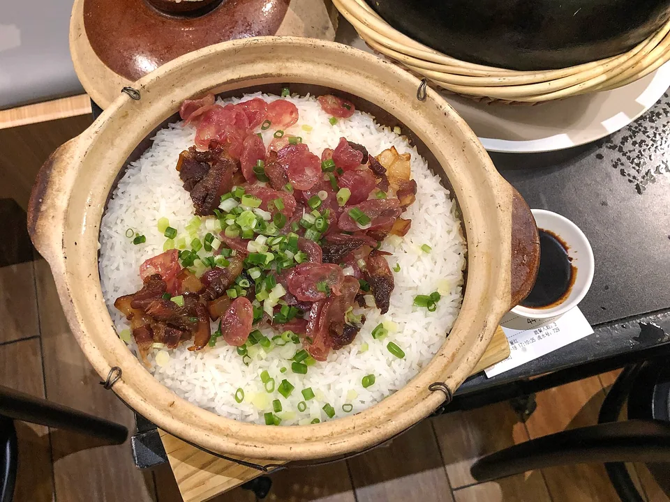 Real finished-dish brief: the clay pot itself or a shallow bowl filled with separate glossy rice grains, sliced red-brown Chinese sausage, cured pork belly, ginger, and chopped scallion; show a distinct golden crisp-rice rim at the bottom edge.