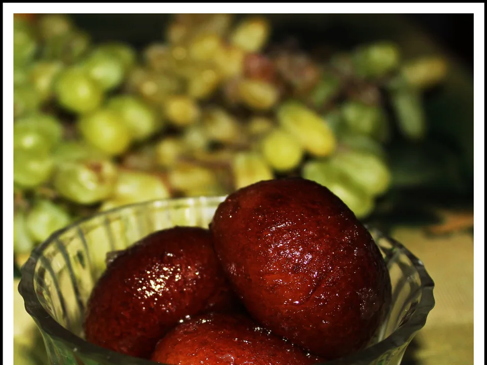 Brown gulab jamun soaked in clear cardamom-rose syrup and garnished with pistachio