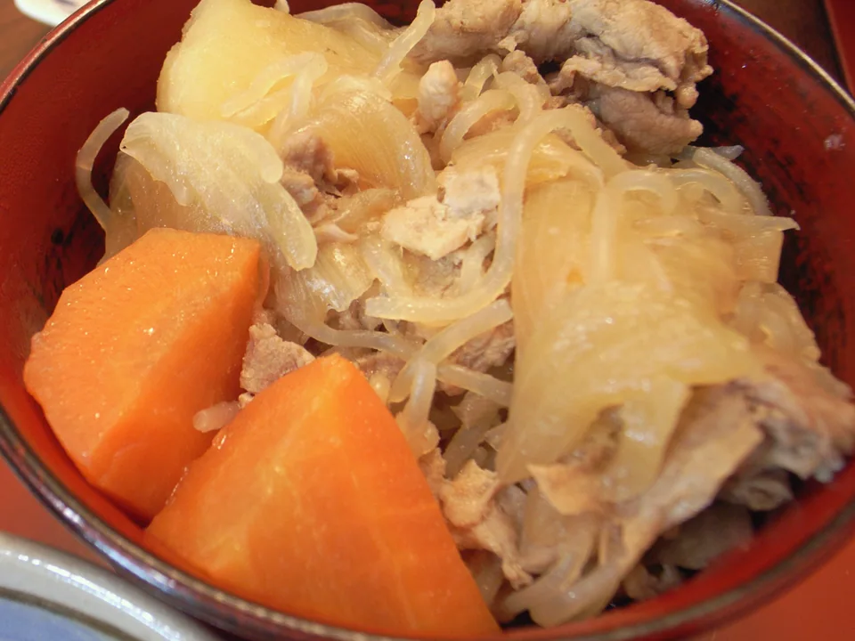 Pork nikujaga in a red bowl with thin pork, chunky potatoes, onion, carrot, and shirataki noodles, tightly cropped away from side dishes