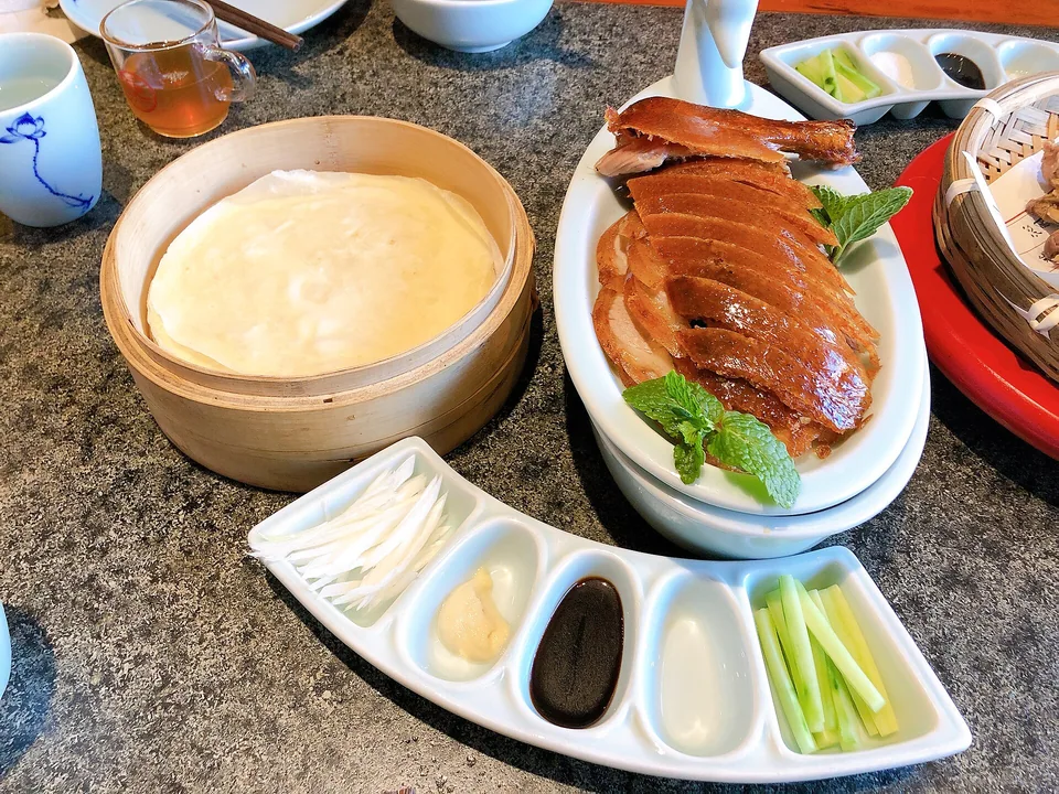Real finished-dish brief: a platter of thin mahogany duck slices with visibly crackled skin, a stack of translucent Mandarin pancakes, cucumber batons, scallion slivers, and an uncovered dish of sweet bean sauce; every wrap component must be visible.