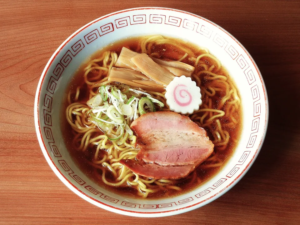 Real photograph of shoyu ramen with clear dark soy broth, yellow wavy noodles, two thick chashu slices, menma, narutomaki and sliced scallions; no egg or nori.