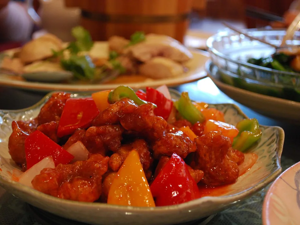 Real finished-dish brief: a shallow white bowl of lacquered golden pork cubes with clearly visible red, yellow, and green bell-pepper squares, onion, ginger, and a few scallion slivers; the red sweet-and-sour sauce should glisten without hiding crisp edges.