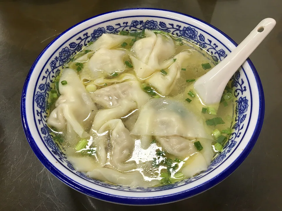 Real finished-dish brief: a deep white bowl with 8–10 plump, semi-translucent wontons in clear golden broth, a whole baby bok choy beside them, ginger threads, and fresh scallion rings floating visibly on top.