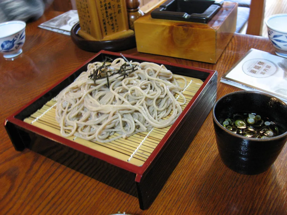Real photograph of chilled soba on a bamboo zaru topped with shredded nori, with separate dark dipping sauce and sliced scallion.